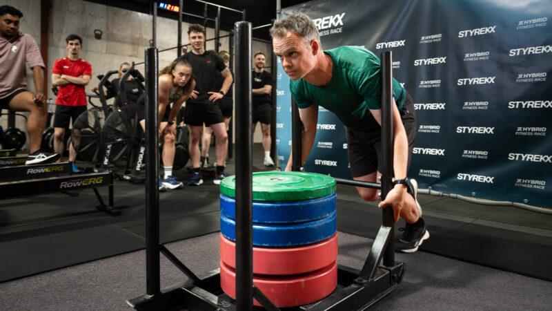 Man duwt zware sled tijdens STYREKX fitnessrace terwijl zijn team gefocust toekijkt in de arena.